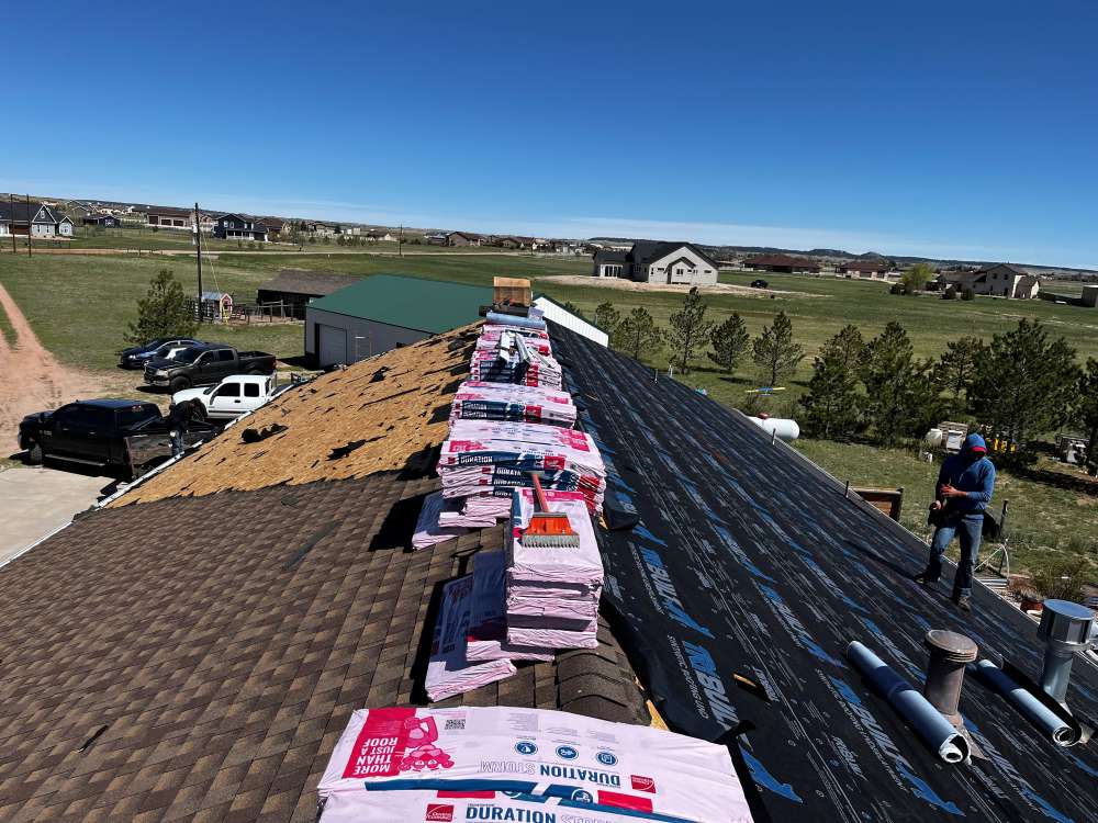 A rooftop under repair with bundles of roofing shingles stacked along the ridge, some protective underlayment installed, and a worker standing on the right. Houses, trees, and vehicles are visible in the background under a clear sky.
