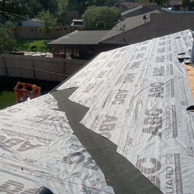A house roof under construction with synthetic underlayment partially installed, some roofing felt in the middle, and construction materials visible on the side. Residential buildings and greenery are in the background.