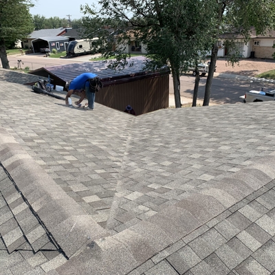 Two people work on a rooftop covered with gray shingles. Trees, a shed, and a street with vehicles are visible in the background on a sunny day.