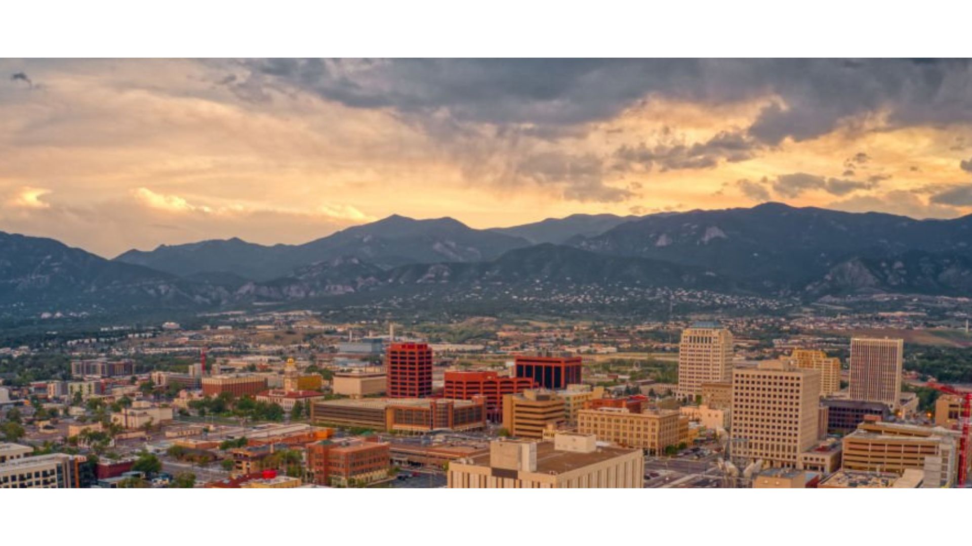 A cityscape at sunset with modern buildings in the foreground and mountains in the background under a cloudy sky.
