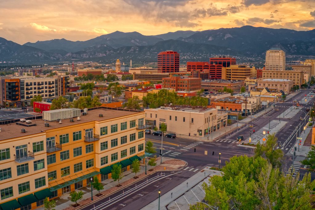 Downtown cityscape at sunset with colorful buildings, empty streets, and distant mountain range under a partly cloudy sky.