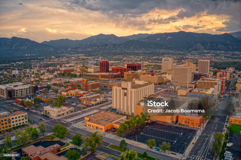Aerial view of downtown Colorado Springs at sunset with buildings, streets, and mountains in the background under a cloudy sky.