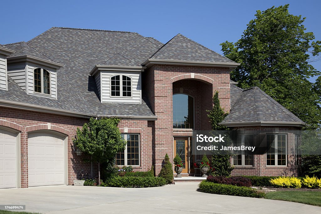 Two-story brick house with a large arched front window, three-car garage, manicured lawn, and shrubs. The sky is clear and blue, and mature trees surround the property.