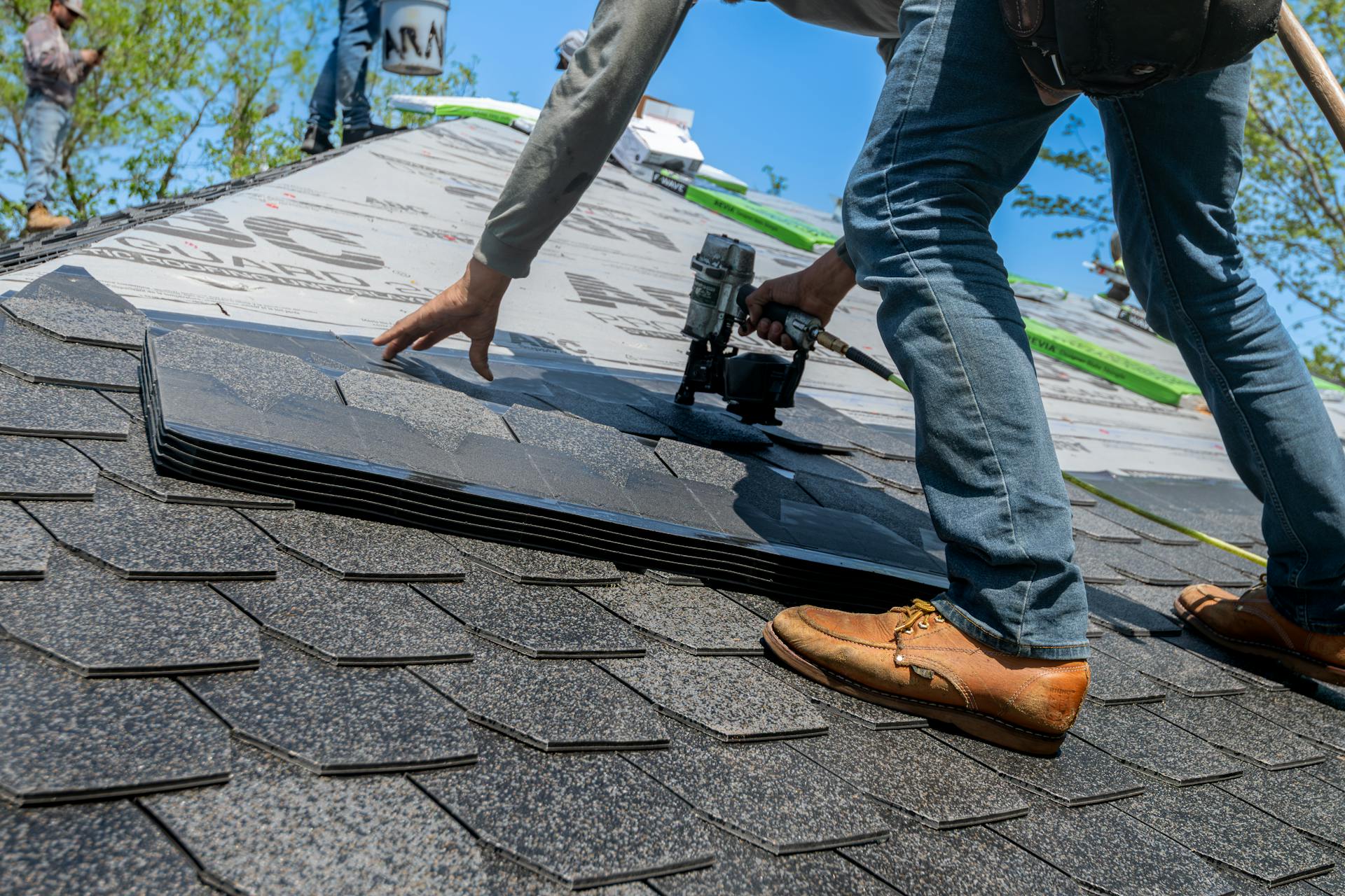 A worker wearing brown shoes and jeans installs asphalt shingles on a roof using a nail gun. The roof is partially covered with underlayment, and the sky is clear in the background.